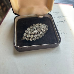 Op de afbeelding: Een zilveren broche met een cluster van heldere strass-steentjes in de vorm van een blad, gepresenteerd in een doos met een fluwelen voering.