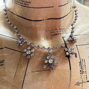 Può includere: Una collana d'argento con una catena delicata e tre ciondoli a forma di fiore adornati con strass trasparenti. La collana è esposta su un manichino di stoffa marrone.