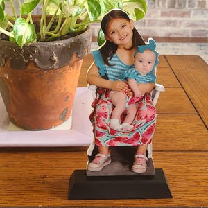 May include: A decorative cutout featuring two young girls. One girl is holding a baby, both seated in a white chair. The baby wears a blue top and the older girl wears a blue striped top. The baby's skirt has a watermelon pattern. The cutout is on a black base.
