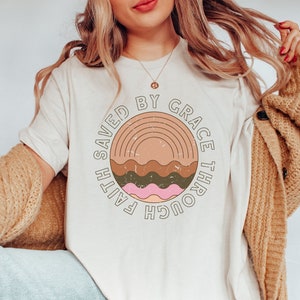 K&ouml;nnte beinhalten: Ein wei&szlig;es T-Shirt mit einem braunen, gr&uuml;nen und rosafarbenen Grafikdesign. Die Grafik zeigt einen Regenbogen und den Text "Faith Saved By Grace Through".
