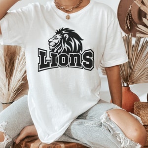 Pode incluir: Uma camiseta branca com um gr&aacute;fico de le&atilde;o preto e a palavra "Lions" em uma fonte preta em negrito.