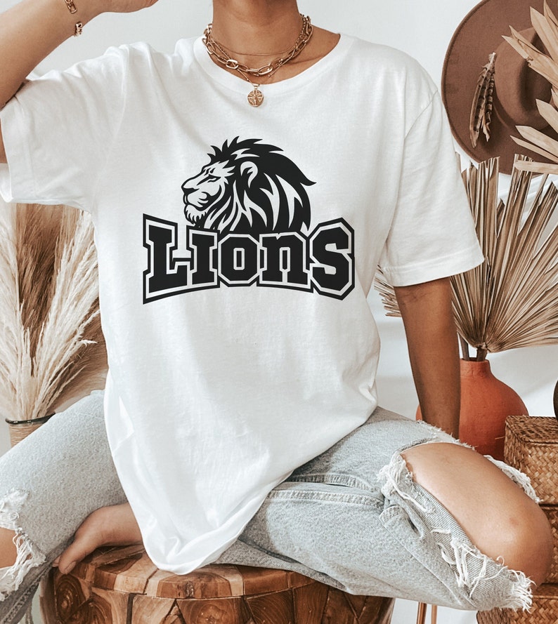 Pode incluir: Uma camiseta branca com um gr&aacute;fico de le&atilde;o preto e a palavra "Lions" em uma fonte preta em negrito.