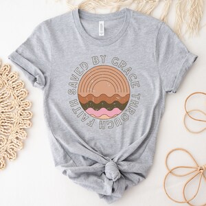 K&ouml;nnte beinhalten: Graues T-Shirt mit einem kreisf&ouml;rmigen Grafikdesign mit dem Text "Gerettet durch Gnade durch Glauben" in einer Vintage-Schriftart. Die Grafik enth&auml;lt auch einen stilisierten Regenbogen und Wellenlinien in Braun-, Rosa- und Oranget&ouml;nen.