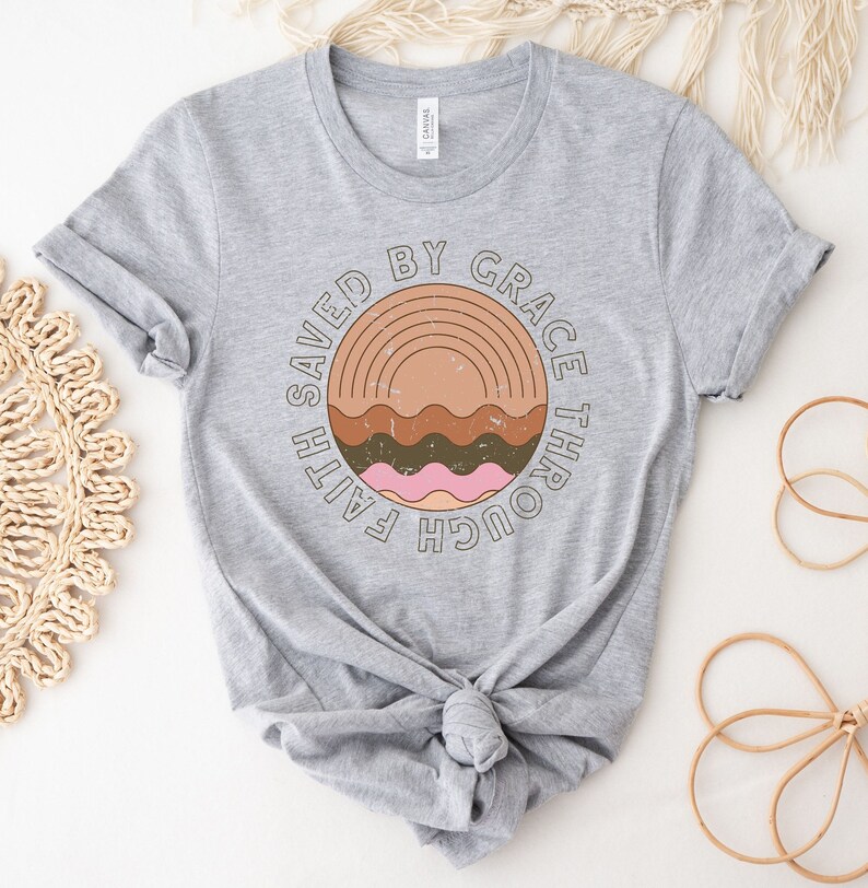 K&ouml;nnte beinhalten: Graues T-Shirt mit einem kreisf&ouml;rmigen Grafikdesign mit dem Text "Gerettet durch Gnade durch Glauben" in einer Vintage-Schriftart. Die Grafik enth&auml;lt auch einen stilisierten Regenbogen und Wellenlinien in Braun-, Rosa- und Oranget&ouml;nen.