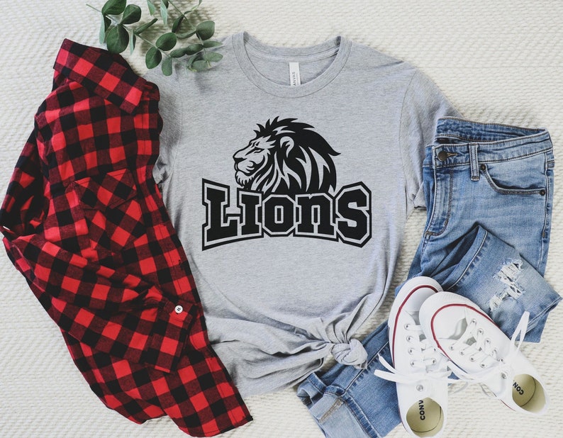 May include: A gray t-shirt with a black lion graphic and the word "Lions" in black text. The shirt is laid out on a white surface with a red and black plaid shirt, blue jeans, and white sneakers.