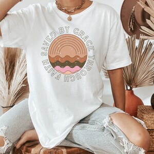 K&ouml;nnte beinhalten: Wei&szlig;es Oversize-T-Shirt mit einem Grafikdesign eines Sonnenuntergangs mit Wellen und dem Text "Saved by Grace Through Faith".