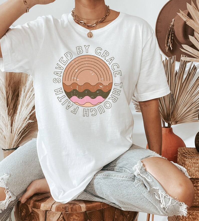 K&ouml;nnte beinhalten: Wei&szlig;es Oversize-T-Shirt mit einem Grafikdesign eines Sonnenuntergangs mit Wellen und dem Text "Saved by Grace Through Faith".