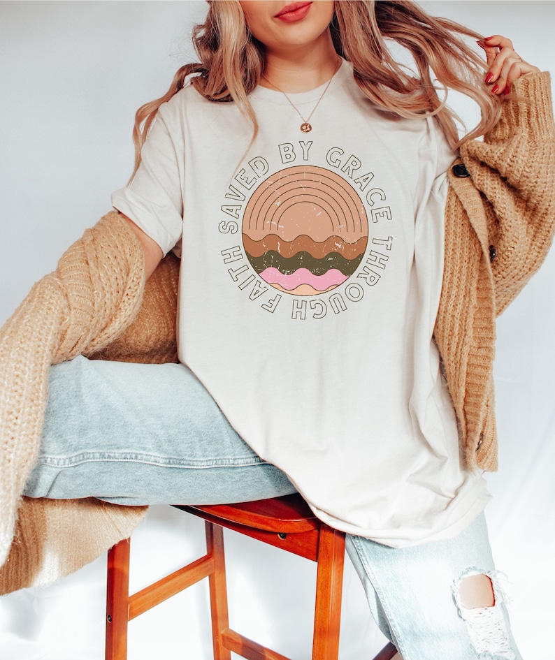 K&ouml;nnte beinhalten: Ein wei&szlig;es T-Shirt mit einem braunen, gr&uuml;nen und rosafarbenen Grafikdesign. Die Grafik zeigt einen Regenbogen und den Text "Faith Saved By Grace Through".