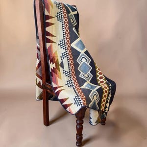 May include: A woven throw blanket featuring a geometric pattern with triangles, diamonds, and chevron shapes in a color palette of navy blue, cream, burgundy, and gold. The blanket is draped over a dark brown wooden chair with turned legs.