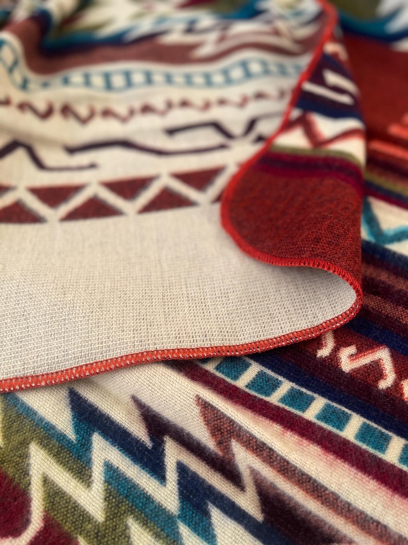 May include: Close-up of a folded blanket with a geometric pattern in shades of red, blue, and cream. The blanket has a red stitched edge. The design includes zigzags, triangles, and other shapes.