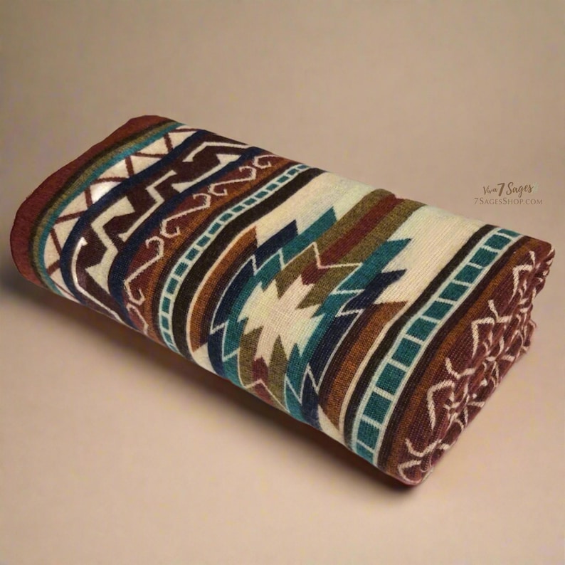 May include: Rolled Southwestern-style blanket with a geometric pattern in shades of brown, cream, teal, and navy blue. The blanket features a repeating design of zigzags, diamonds, and other shapes. The edges are finished with a dark brown border.