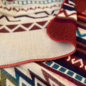 K&ouml;nnte beinhalten: Nahaufnahme einer gefalteten Decke mit einem geometrischen Muster in Rot-, Blau- und Cremet&ouml;nen. Die Decke hat einen rot gen&auml;hten Rand. Das Design zeigt Zickzacklinien, Dreiecke und andere Formen.