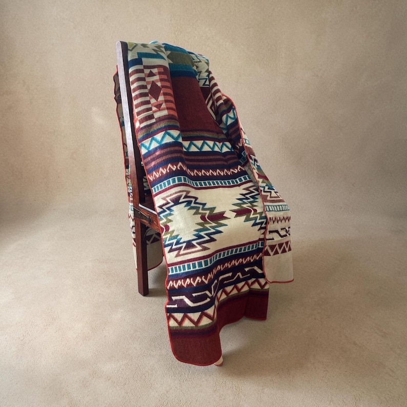 May include: A colorful woven blanket with a geometric pattern draped over a wooden chair. The blanket features a cream-colored background with red, blue, teal, and purple geometric designs. The blanket's edges are trimmed in a deep red color.