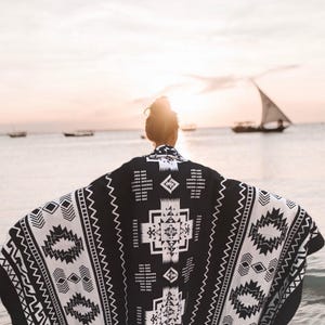 Peut inclure: Une couverture de plage à motifs géométriques noirs et blancs. La couverture est drapé sur une personne debout sur une plage avec l'océan en arrière-plan.