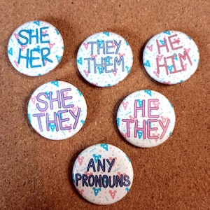 Può includere: Una collezione di sei bottoni rotondi con pronomi. Ogni bottone presenta uno sfondo bianco con un motivo di simboli di genere rosa e blu. I bottoni mostrano pronomi come "She/Her", "They/Them", "He/Him", "She/They", "He/They" e "Any Pronouns" in vari colori.