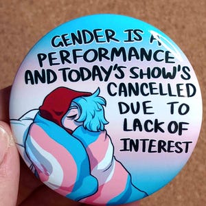 May include: A circular badge with a blue and pink gradient background. The text reads "GENDER IS A PERFORMANCE AND TODAY'S SHOW'S CANCELLED DUE TO LACK OF INTEREST." An illustration of two figures embracing is in the center.