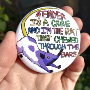 Puede incluir: Un botón redondo y blanco con el texto "GENDER IS A CAGE AND I'M THE RAT THAT CHEWED THROUGH THE BARS" en varios colores. Una ilustración de rata morada y blanca aparece en el botón.