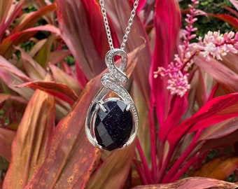 May include: A silver necklace with a dark blue gemstone pendant. The pendant is set in a silver frame with a swirl design and small clear stones. The necklace is photographed against a backdrop of red and green foliage.