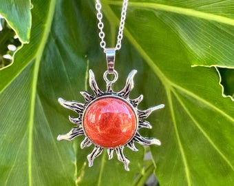 May include: A silver-toned necklace featuring a sun-shaped pendant with a central, round, reddish-orange stone. The pendant has radiating, flame-like metal rays extending from the center, suspended from a delicate silver-colored chain.