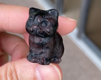 Könnte beinhalten: Eine kleine, schwarze und violette Katzenfigur aus Edelstein geschnitzt. Die Figur sitzt mit den Pfoten unter sich.
