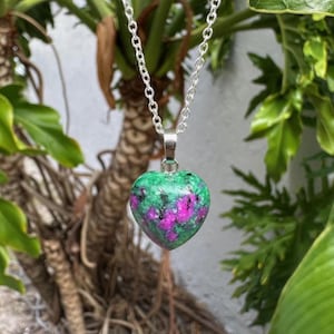 May include: A heart-shaped pendant necklace with a silver chain. The pendant is made of a green and pink stone. The necklace is displayed against a backdrop of green foliage.