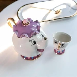 Może przedstawiać: Biały ceramiczny zestaw do herbaty z motywem z kreskówki. Czajniczek ma fioletową pokrywkę z różowym klejnotem, a filiżanka ma złoty uchwyt. Zestaw ma złote wykończenia i dekoracyjną podstawę.