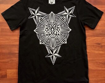 Op de afbeelding: Zwart T-shirt met een wit grafisch ontwerp met een gestileerde zon met acht punten en drie sterren. Het ontwerp is gecentreerd op het shirt.