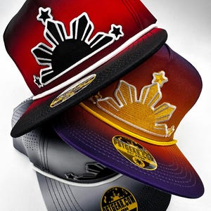 Könnte beinhalten: Drei Ombre-Baseballkappen mit einem Sonnen- und Sternendesign. Die Kappen sind rot, lila und grau mit weißen Nähten. Das Sonnen- und Sternendesign ist schwarz auf der roten Kappe, gold auf der lila Kappe und weiß auf der grauen Kappe. Jede Kappe hat ein schwarzes und gelbes Etikett mit dem Text "PSTGEAR.COM EST.2006".