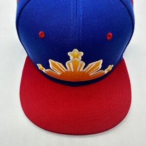 Op de afbeelding: Een blauwe en rode baseballpet met een geborduurd geel zon- en sterrendesign. De zon heeft een rood en oranje verloop.