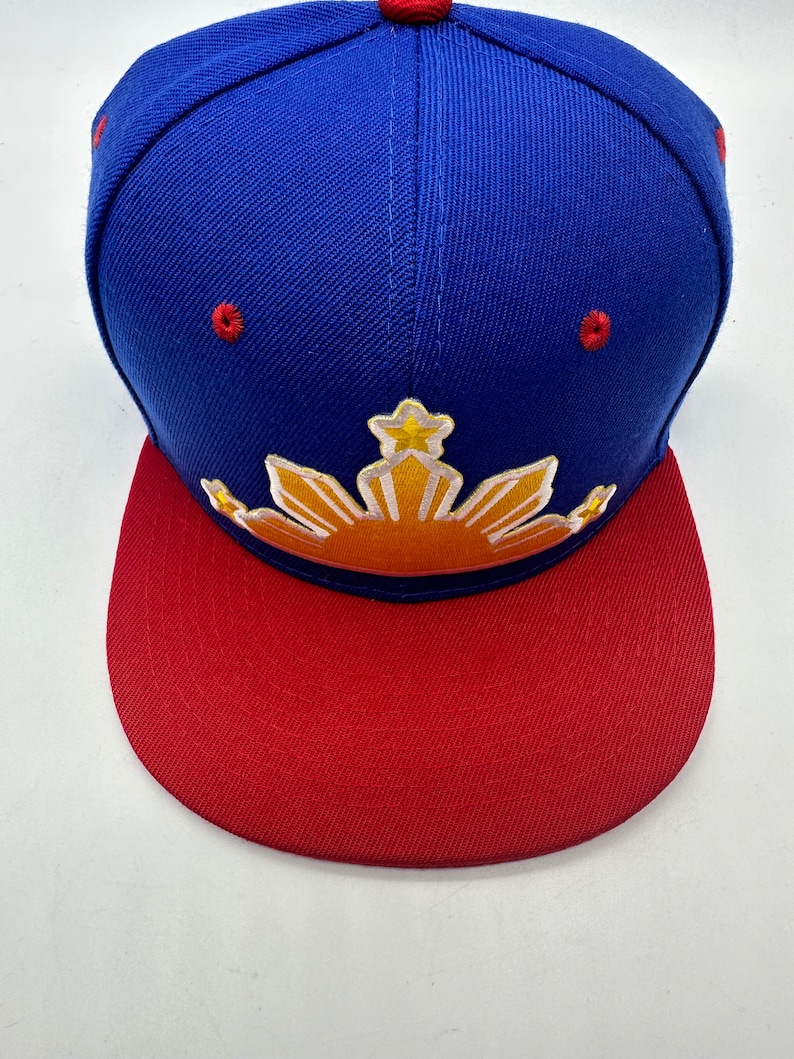 Op de afbeelding: Een blauwe en rode baseballpet met een geborduurd geel zon- en sterrendesign. De zon heeft een rood en oranje verloop.
