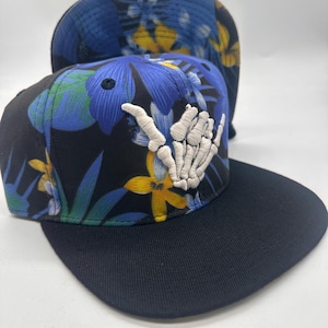 May include: A black snapback cap featuring a vibrant floral pattern in shades of blue, green, and yellow. The front panel displays a prominent white embroidered skeleton hand making a shaka gesture. The cap has a solid black flat brim.