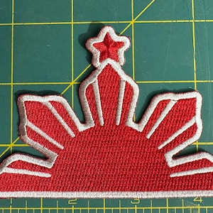 May include: Embroidered patch featuring a red sunburst design with eight rays and three five-pointed stars. The patch has a white border and is likely for clothing or accessories.