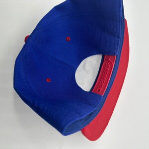 Op de afbeelding: Een blauwe en rode baseballpet met een rode onderkant. De pet heeft een drukknopsluiting en een klein gaatje bovenop.