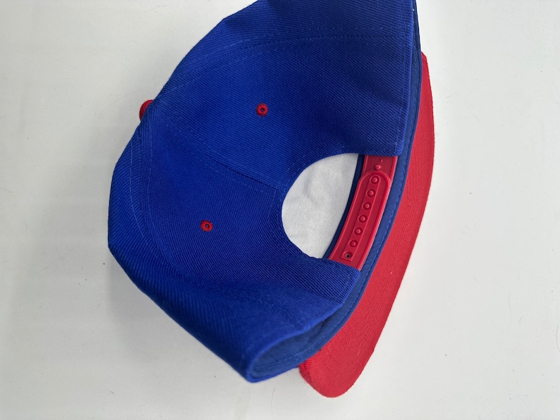 Op de afbeelding: Een blauwe en rode baseballpet met een rode onderkant. De pet heeft een drukknopsluiting en een klein gaatje bovenop.