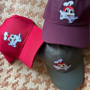 May include: Three baseball caps featuring a cartoon character wearing a chef's hat and making a shaka sign. The caps are red, burgundy, and olive green.