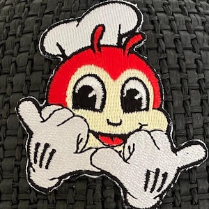 May include: A red and white cartoon bee with a chef's hat, wearing white gloves and giving a shaka sign. The bee has large eyes and a smiling mouth.