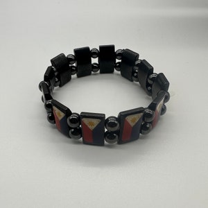 Puede incluir: Un brazalete negro con peque&ntilde;as cuentas negras y abalorios rectangulares con la bandera de Filipinas. La bandera es roja, blanca y azul con un sol amarillo en el centro.