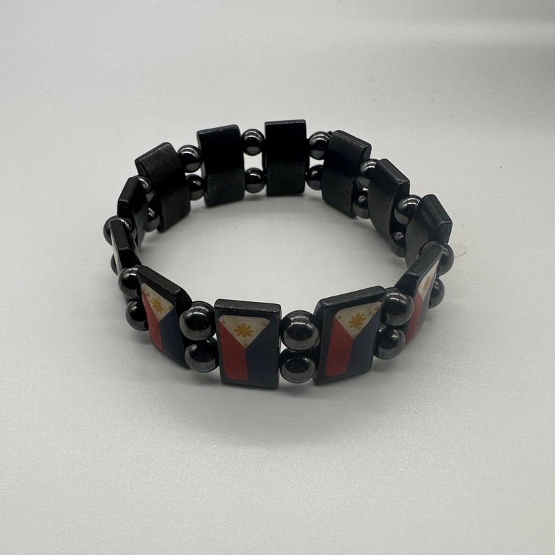 Puede incluir: Un brazalete negro con peque&ntilde;as cuentas negras y abalorios rectangulares con la bandera de Filipinas. La bandera es roja, blanca y azul con un sol amarillo en el centro.