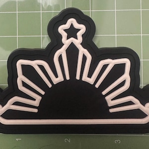 Puede incluir: Un emblema en blanco y negro con un diseño de sol con tres estrellas. El emblema tiene un fondo negro con un contorno y detalles blancos. El sol tiene forma semicircular con rayos que se extienden hacia afuera.