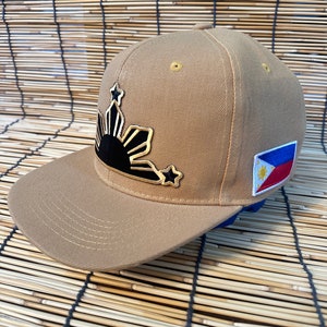 Könnte beinhalten: Eine beige Basecap mit einem gestickten Sonnen- und Sternenmotiv in Schwarz und Gold. Die Kappe hat außerdem einen kleinen philippinischen Flaggen-Patch an der Seite.