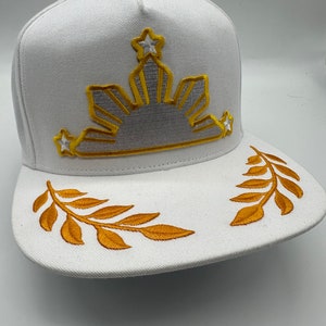Puede incluir: Gorra de béisbol blanca con un diseño de sol y estrellas bordado en oro en la parte delantera y hojas bordadas en oro en la visera.