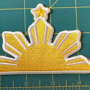 May include: Embroidered patch with a yellow sunburst design, white edging, and three yellow stars. The rectangular patch has a detailed, textured appearance, ideal for application on clothing or accessories.
