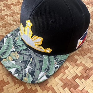 May include: A black and green baseball cap with a white and yellow sun and stars design on the front. The back of the cap has a small Philippine flag.