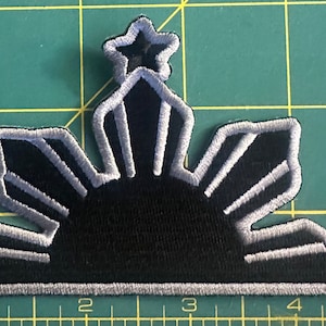 May include: A black and silver embroidered patch featuring the sun and three stars, a symbol of the Philippines.