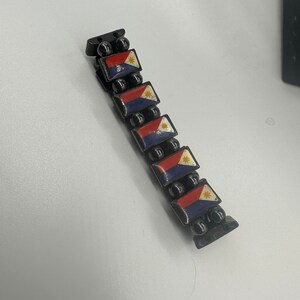 Puede incluir: Un brazalete negro con peque&ntilde;as cuentas negras y abalorios rectangulares con la bandera de Filipinas. Los abalorios son rojos, blancos y azules con una estrella amarilla.