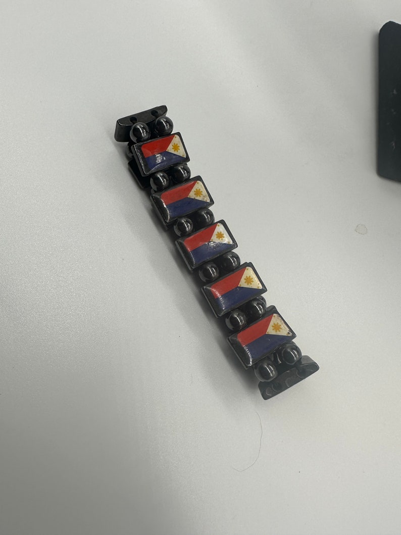 Puede incluir: Un brazalete negro con peque&ntilde;as cuentas negras y abalorios rectangulares con la bandera de Filipinas. Los abalorios son rojos, blancos y azules con una estrella amarilla.