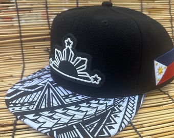 May include: Black baseball cap featuring a white and black patterned brim. The cap has a white emblem of a sun with rays and stars. A small Philippine flag is on the side.