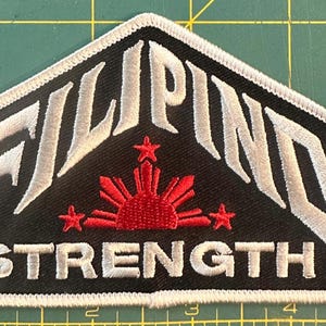 May include: Embroidered patch with the word "FILIPINO" in white, arched over "STRENGTH" in white, on a black background. A red sunburst design with three stars is centered below "FILIPINO". The patch has a white border.