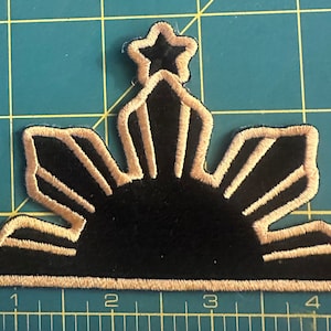 May include: A black and gold embroidered patch featuring the sun and three stars, a symbol of the Philippines.