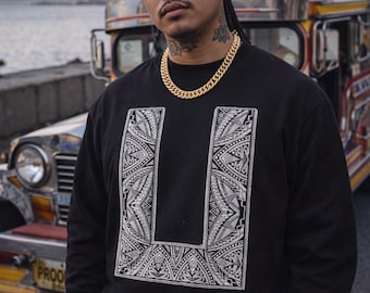 Op de afbeelding: Een zwart sweatshirt met een wit tribaal ontwerp en een gouden ketting. De achtergrond bevat een kleurrijke jeepney en een skyline.